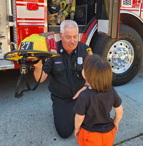 27s about page Firefighter from 27s kneeling and handing his helmet to a little girl facing him. She is wearing a black shirt and orange pants. A fire apparatus with its door open is the backgroud.